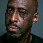 A close-up portrait of a man shedding a tear against a gray background, depicting sadness and contemplation.